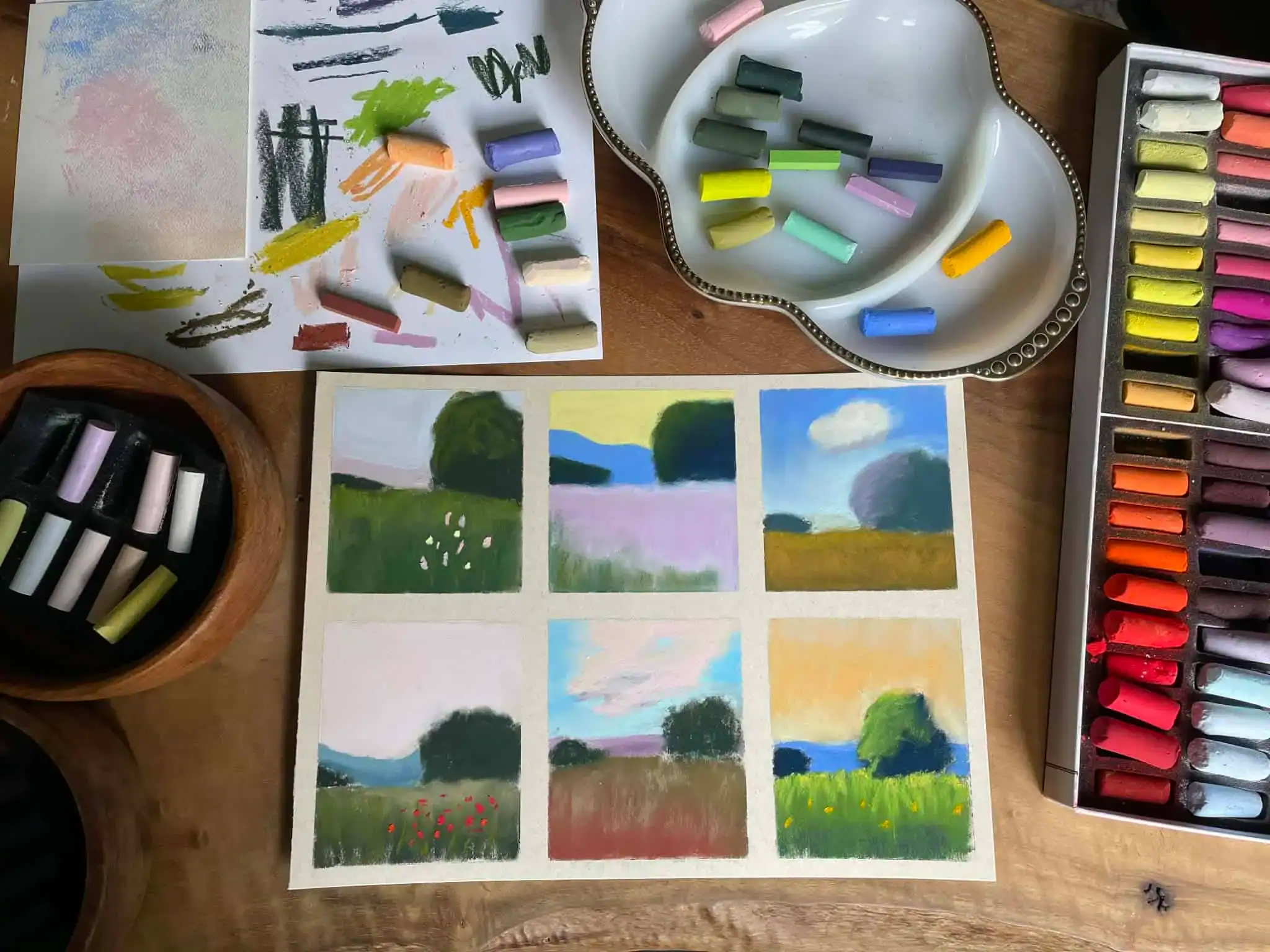 Overhead view of an open box of soft pastels sorted by color, beside a dish of pastel pieces and a sheet of small landscape studies.