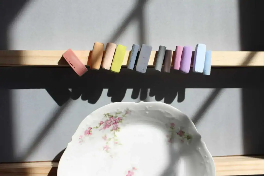 Valerie McKeehan's soft pastels lined up on a wooden shelf in window light, beside a floral porcelain plate.