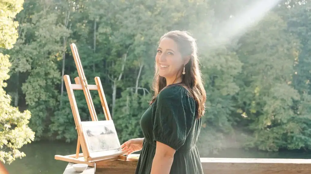 Valerie McKeehan smiling beside her easel on a sunlit deck above a wooded lake, with a small soft pastel painting in progress.