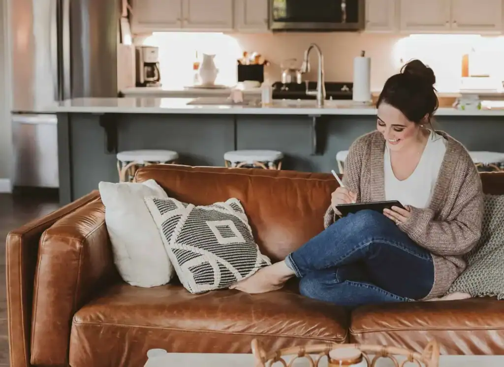 Valerie McKeehan sketching on a tablet while seated on a brown leather sofa in a bright, warm living room.